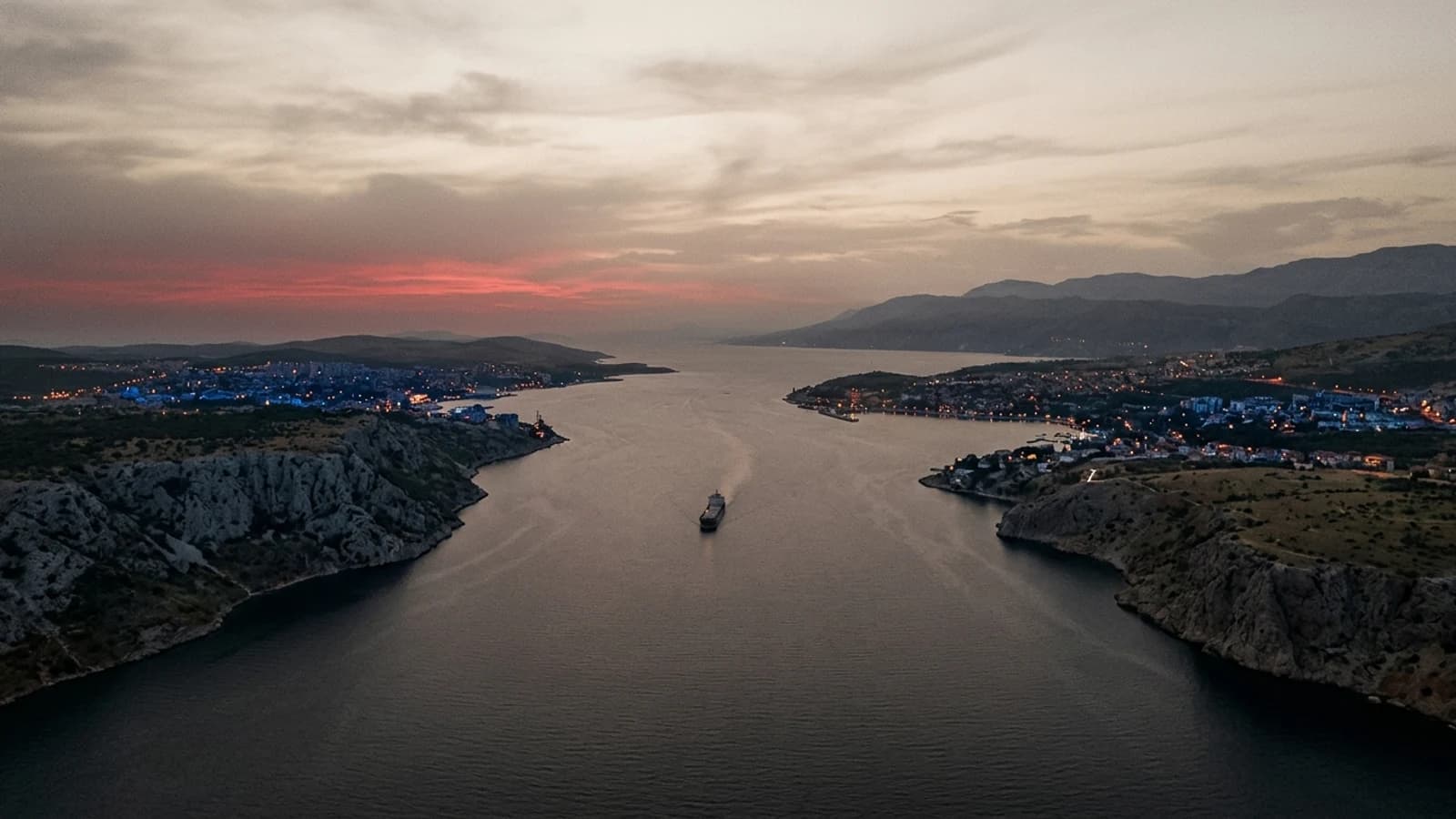 A narrow sea strait at dusk seen from above, a single cargo vessel passing between two shores as city lights appear.