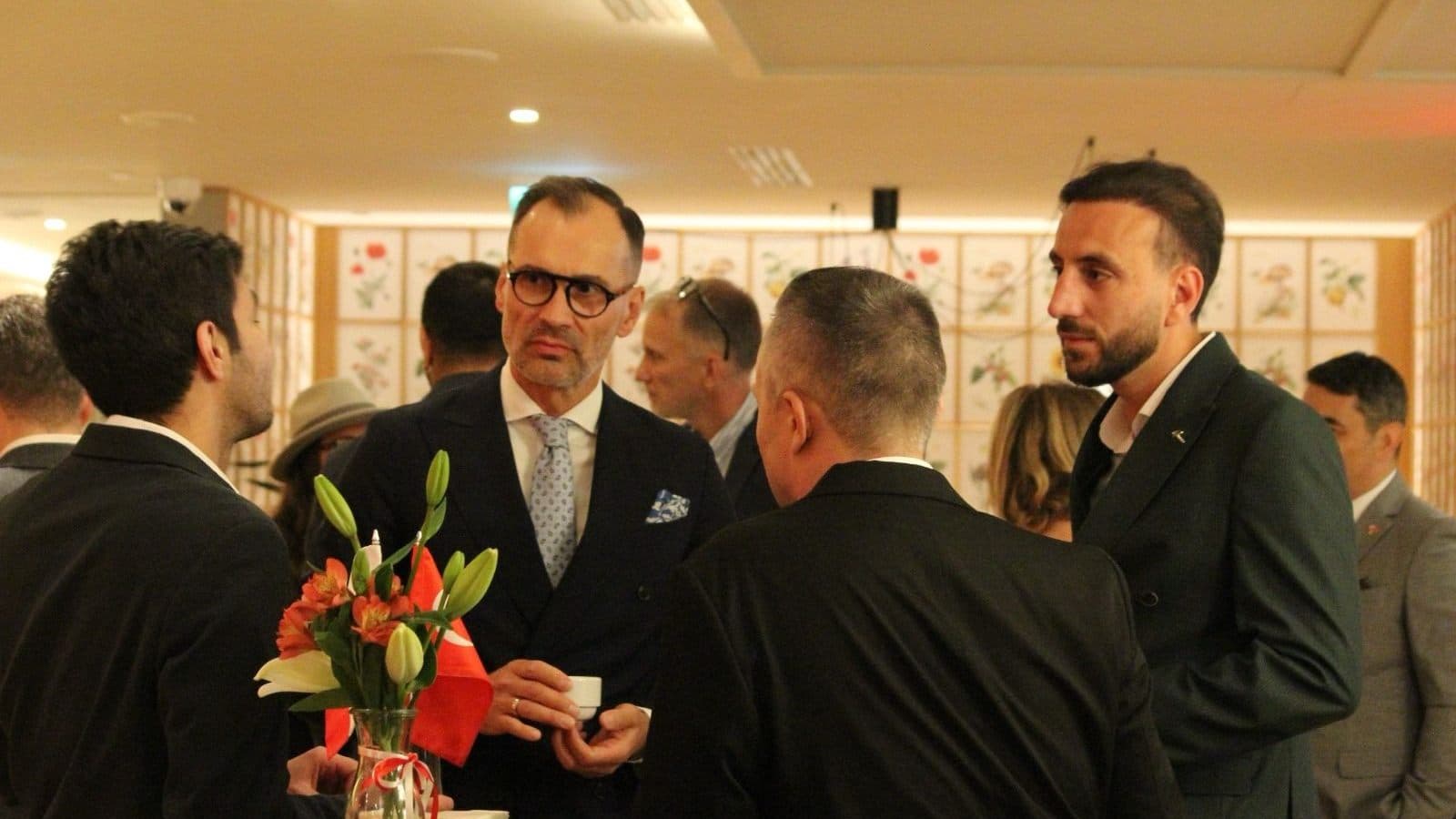Guests standing in small groups in conversation at an evening reception, with Turkish coffee being served.